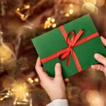 Ruky držiace elegantne zabalený zelený darčekový poukaz s červenou stužkou na pozadí rozsvieteného vianočného stromčeka.